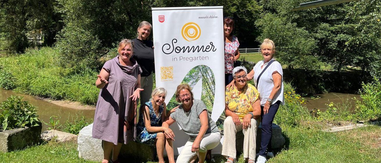 Fünf Frauen posieren vor einem Banner mit der Aufschrift 'Sommer in Pregarten'. Sie sitzen und stehen auf einem Rasen mit einem Fluss und Bäumen im Hintergrund.