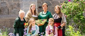 Eine Gruppe von Frauen und Mädchen in historischer Kleidung posiert für ein Foto im Garten, hält Blumen und lächelt.