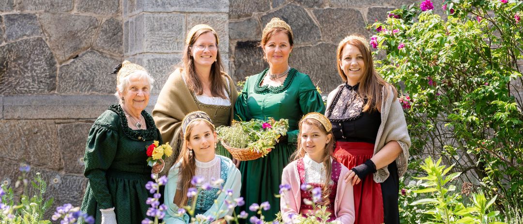 Eine Gruppe von Frauen und Mädchen in historischer Kleidung posiert für ein Foto im Garten, hält Blumen und lächelt.