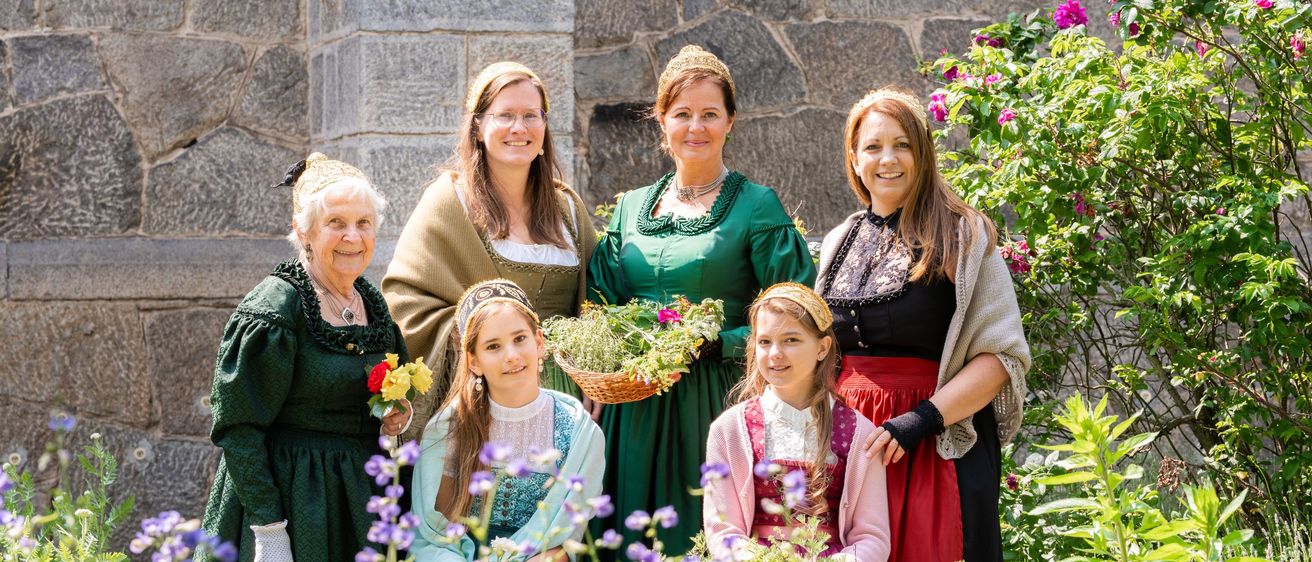 Eine Gruppe von Frauen und Mädchen in historischer Kleidung posiert für ein Foto im Garten, hält Blumen und lächelt.