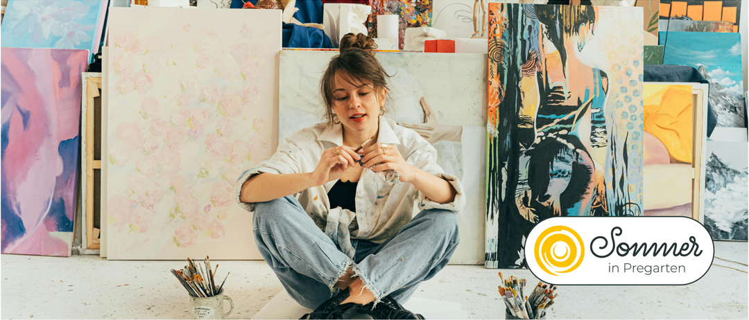 Eine Frau sitzt auf dem Boden in einem Kunstatelier, hält einen Pinsel in der Hand. Sie trägt ein weißes Hemd und blaue Jeans, umgeben von Leinwänden und Kunstzubehör.