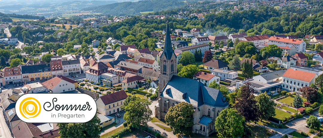 Luftaufnahme einer Stadt mit einer großen Kirche in der Mitte, umgeben von verschiedenen Gebäuden und üppiger Grünfläche. Die Kirche hat einen hohen Turm und eine Uhr an der Seite.