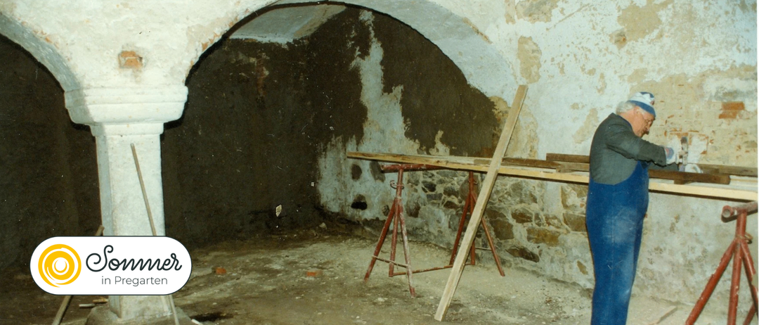 Ein unfertiger Keller mit Steinmauern und einem roten Metallständer, der eine Holzplanke hält, zeigt abblätternden Putz und eine raue Umgebung.
