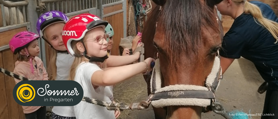 Ein junges Mädchen mit rotem Helm und Brille streichelt ein Pferd. Ein weiteres Mädchen mit grünem Helm hält einen rosa Gegenstand. Sie sind in einer Scheune.