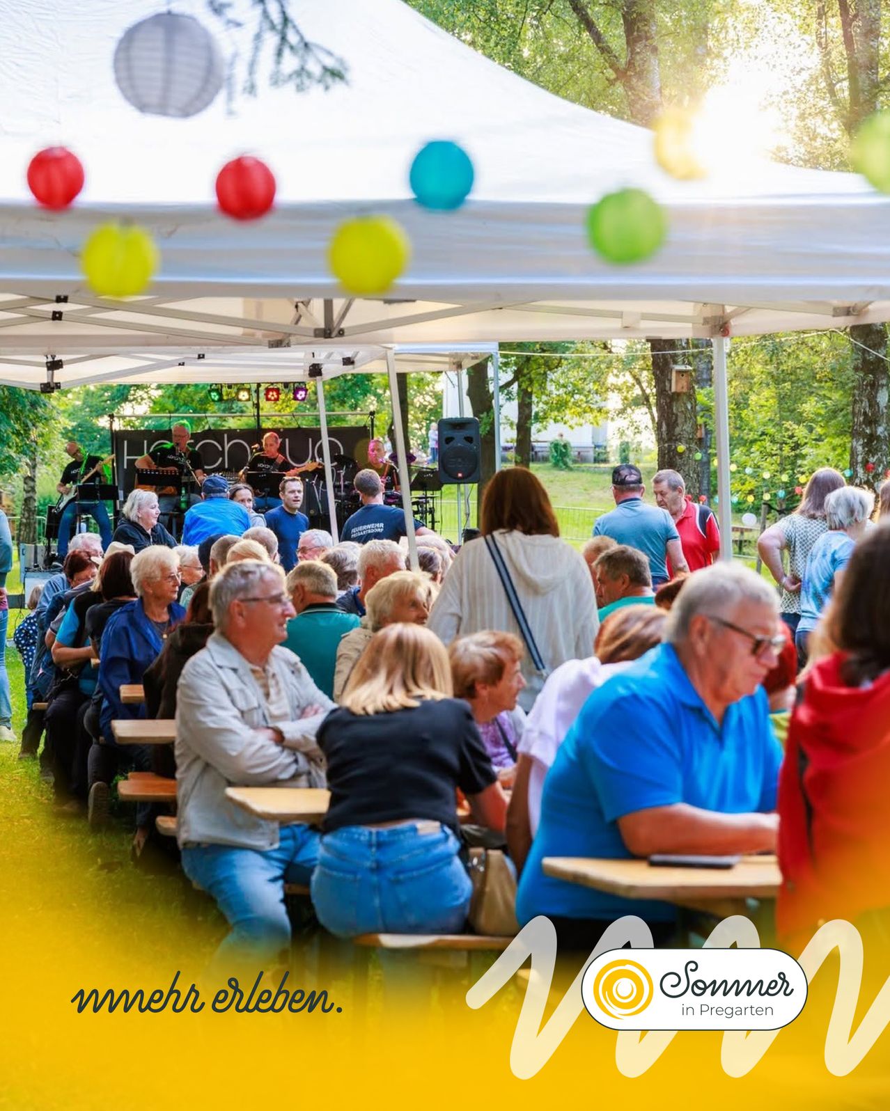 Eine Gruppe von Menschen versammelt sich unter einem Zelt für eine Outdoor-Veranstaltung. Eine Band spielt auf der Bühne mit einem Lautsprecher. Das Publikum sitzt an Tischen, einige stehen und beobachten. Die Umgebung ist im Freien mit Bäumen und einer Rasenfläche.