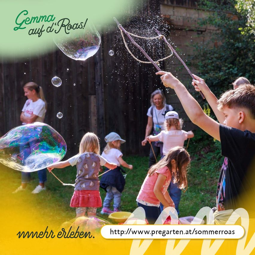 Kinder spielen im Garten und erzeugen mit Stäben Seifenblasen. Eine große Blase schwebt im Vordergrund. Der Hintergrund zeigt einen Holzzaun und Grünpflanzen.