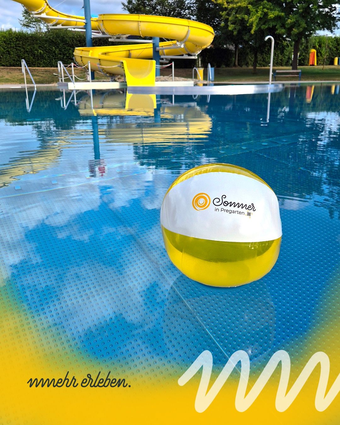 Ein gelber und weißer Strandball mit den Worten 'Sommer in Pregarten' schwimmt in einem blau-gelben Pool mit einem gelben Wasserrutsche im Hintergrund.