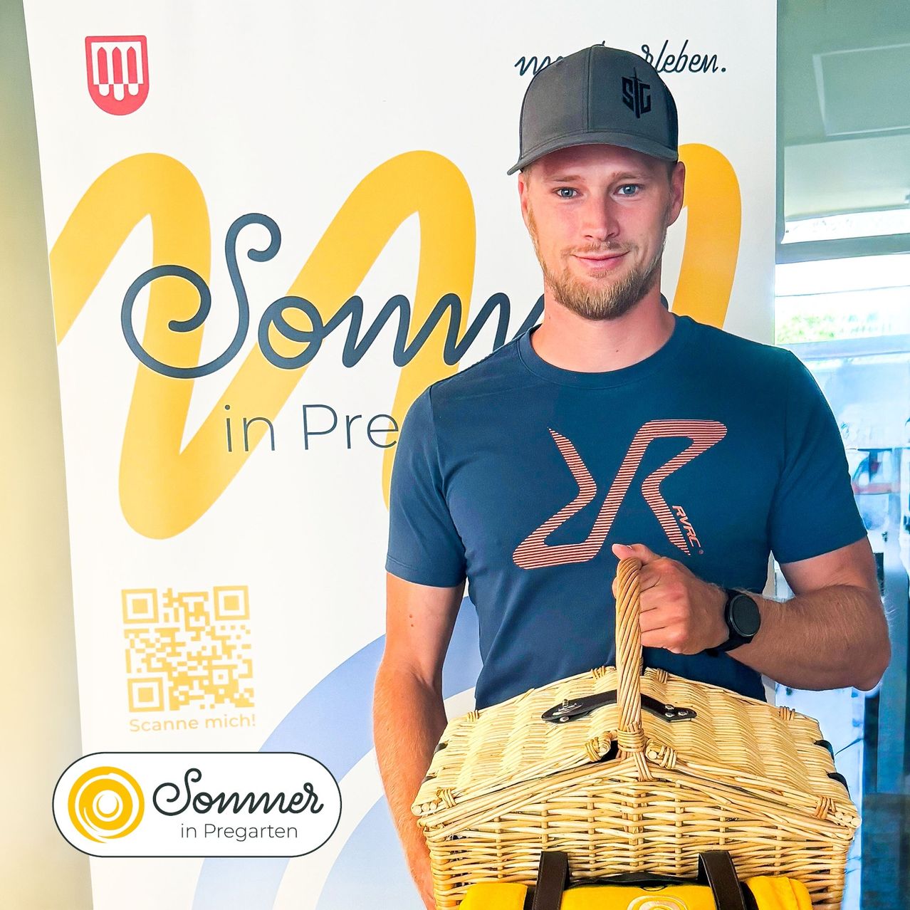 Ein Mann mit einem blauen T-Shirt mit rotem Logo und einer schwarzen Kappe hält einen Weidenkorb vor einem Banner, das 'Sommer in Pre' steht.
