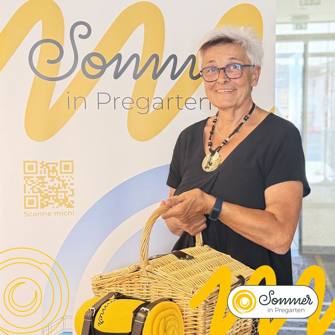 An older woman with white hair wearing glasses and a necklace is standing in front of a banner that says 'Sommer in Pregarten'. She is holding a wicker basket with a yellow blanket inside.