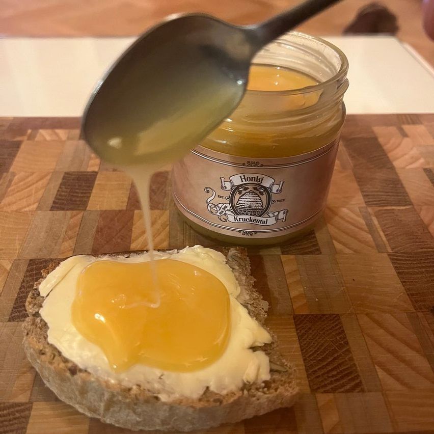 Ein Löffel gießt Honig auf ein Stück Brot mit Butter auf einem Holzschneidbrett. Ein Glas Honig ist in der Nähe.