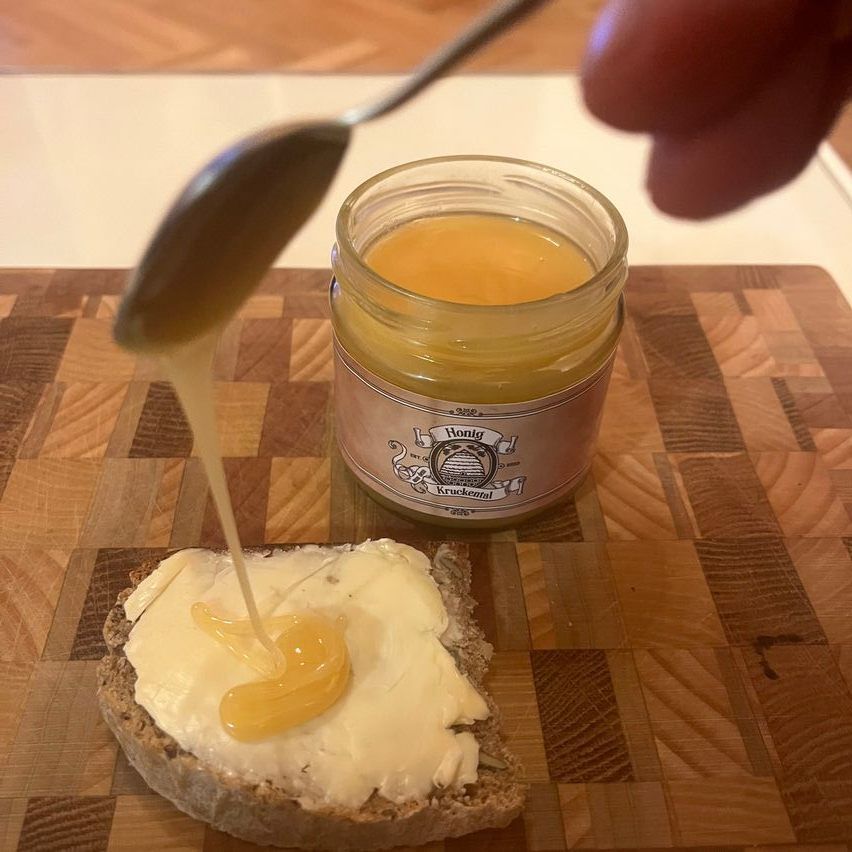 Eine Person benutzt einen Löffel, um Honig auf ein Stück Brot zu geben. Ein Glas Honig steht auf einem hölzernen Schneidebrett in der Nähe.