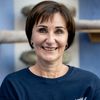 Eine Frau mit kurzen braunen Haaren und blauen Augen lächelt und trägt ein blaues T-Shirt mit weißem Text. Sie steht vor einer blauen Wand mit einem Holzregal.