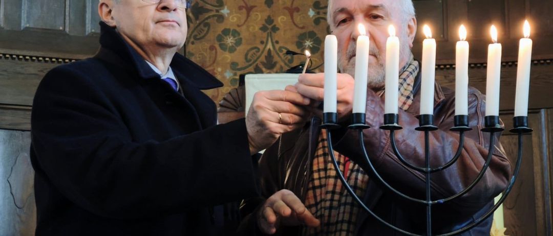 Zwei Männer zünden Kerzen an einer Menora in einer Kirche an. Einer hält eine Kerze, während der andere sie anzündet.
