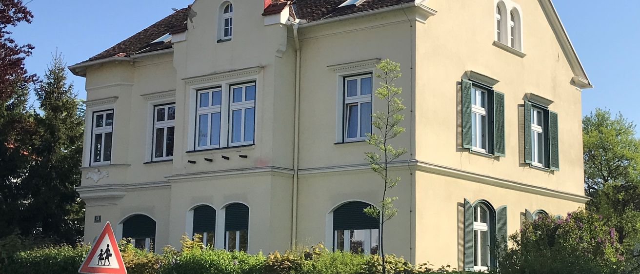 Ein großes beiges Haus mit einem braunen Dach und grünen Fensterläden, das einen Kreuz auf der Spitze und mehrere Fenster hat. Ein Busch und ein Holzzaun stehen vor dem Haus.