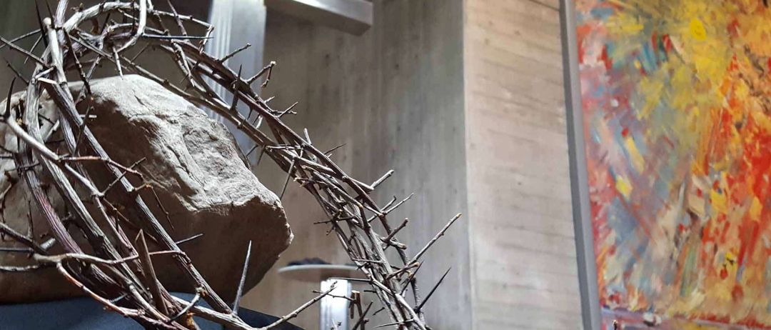 Eine Dornenkrone ruht auf einem Felsen vor einem Kreuz in einer Kirche. An der Wand befindet sich ein farbenfrohes Gemälde.