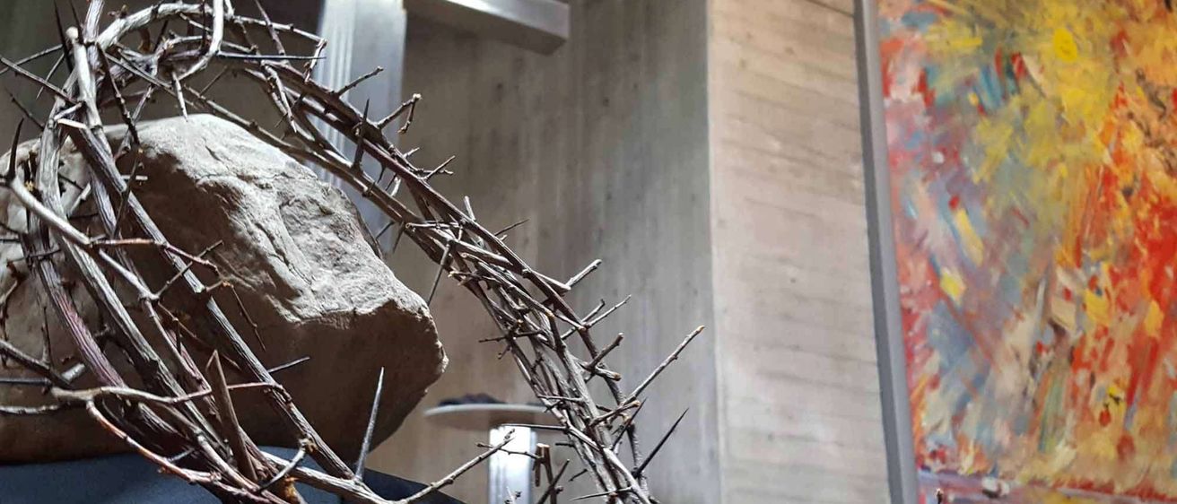 Eine Dornenkrone ruht auf einem Felsen vor einem Kreuz in einer Kirche. An der Wand befindet sich ein farbenfrohes Gemälde.