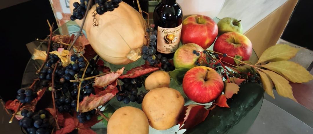 Ein Glastisch zeigt ein Erntearrangement mit Kürbissen, Äpfeln, Trauben und Blättern. Zwei Flaschen, eine beschriftet, stehen ebenfalls auf dem Tisch.
