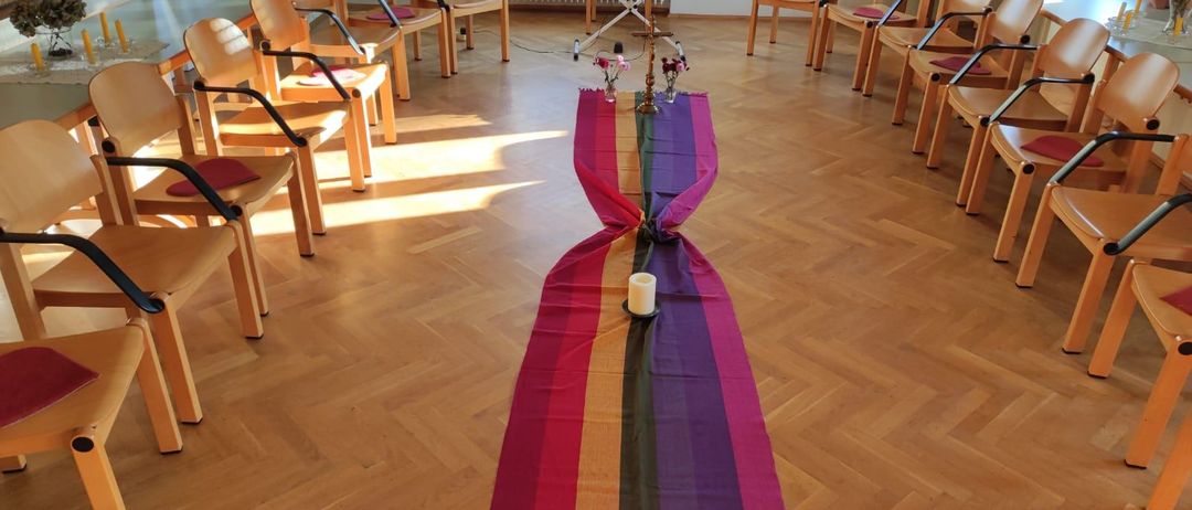 Ein leerer Raum mit hölzernen Stühlen, die im Kreis angeordnet sind. Ein farbenfroher Teppich liegt auf dem Boden mit einer Kerze in der Mitte. Ein Heizkörper steht an der Wand. Ein Tisch und Stühle sind auf der linken Seite. Ein Gemälde hängt an der Wand.