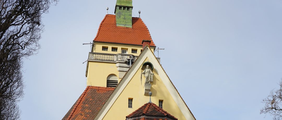 Eine gelbe Kirche mit roten Dächern steht in einer Stadt. Sie hat einen Turm mit grüner Spitze und eine Statue auf der Vorderseite. Es gibt zwei große Türen und mehrere Fenster. Vor der Kirche steht ein kleiner Brunnen.