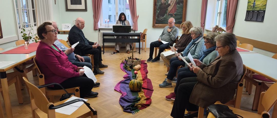 Eine Gruppe älterer Menschen sitzt in einem Kreis und liest von Papieren. Eine Frau steht in der Nähe eines Fensters, und ein Gemälde von Jesus ist an der Wand.