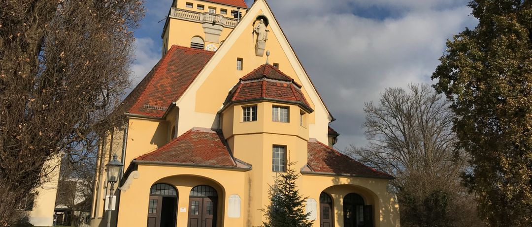 Eine große gelbe Kirche mit roten Dächern, mit einer Statue auf ihrer Fassade, steht unter einem bewölkten Himmel. Um die Kirche herum befinden sich Bäume und Sträucher, und sie hat mehrere gewölbte Eingänge.