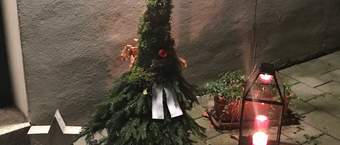 Ein Weihnachtsbaum in Form eines gebogenen Zweiges mit einem roten Ball oben und einer Schleife, neben einer Laterne mit brennenden Kerzen und einem Stern.