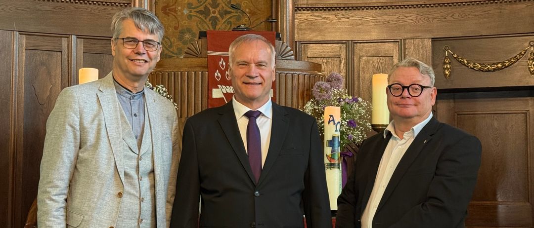 Drei Männer stehen vor einem Kirchenaltar. Ein Mann trägt eine Weste, einer einen Anzug mit Krawatte und der letzte einen Anzug mit weißem Hemd.