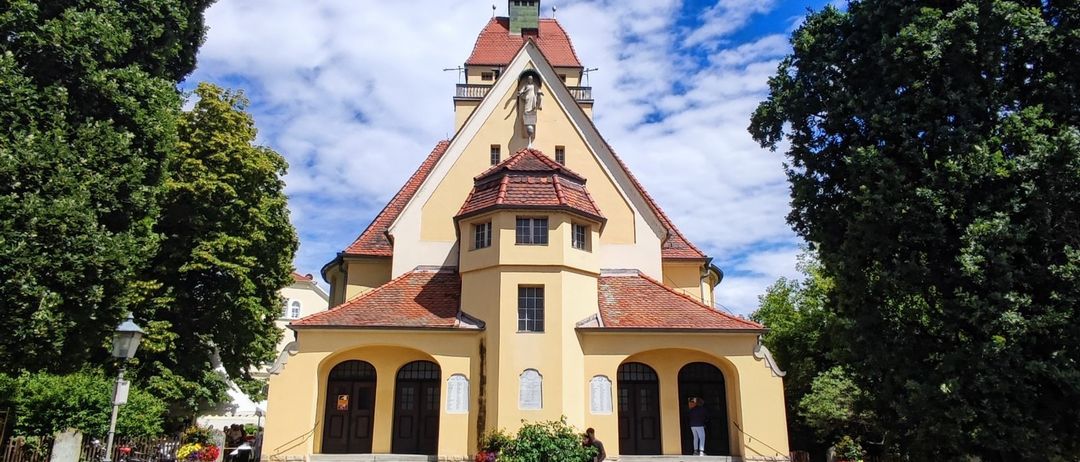 Eine gelbe Kirche mit roten Dächern und großen Rundbogenfenstern. Eine Statue schmückt die Spitze und Menschen betreten durch die Vordertür.