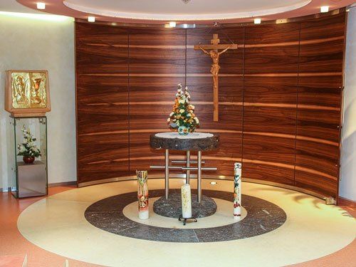 Eine kleine Kapelle mit einem hölzernen Altar und einem Kreuz an der Wand. Eine Blumenarrangement und Kerzen stehen auf dem Altar.