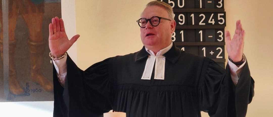 Ein Mann in einem schwarzen Gewand steht an einem kerzenbeleuchteten Altar und erhebt seine Hände zum Gebet. Hinter ihm sind ein großes Gemälde und eine Anzeigetafel zu sehen.