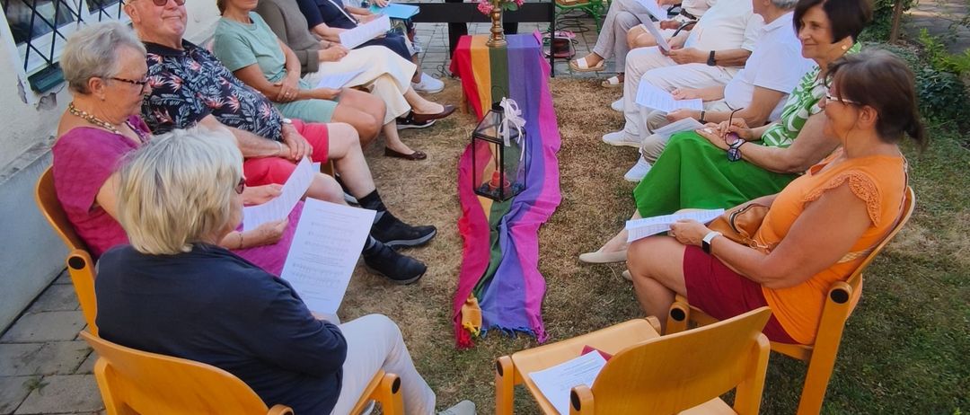 Eine Gruppe älterer Menschen sitzt auf Stühlen auf einem Rasen, liest Papiere und hört einem Mann zu, der vor einem Klavier steht.