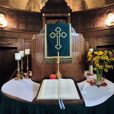 Ein Altar mit einem Kreuzbanner, einem offenen Buch, einer Kerze und Blumen auf einem Holztisch. Der Altar ist von Holzwänden umgeben.