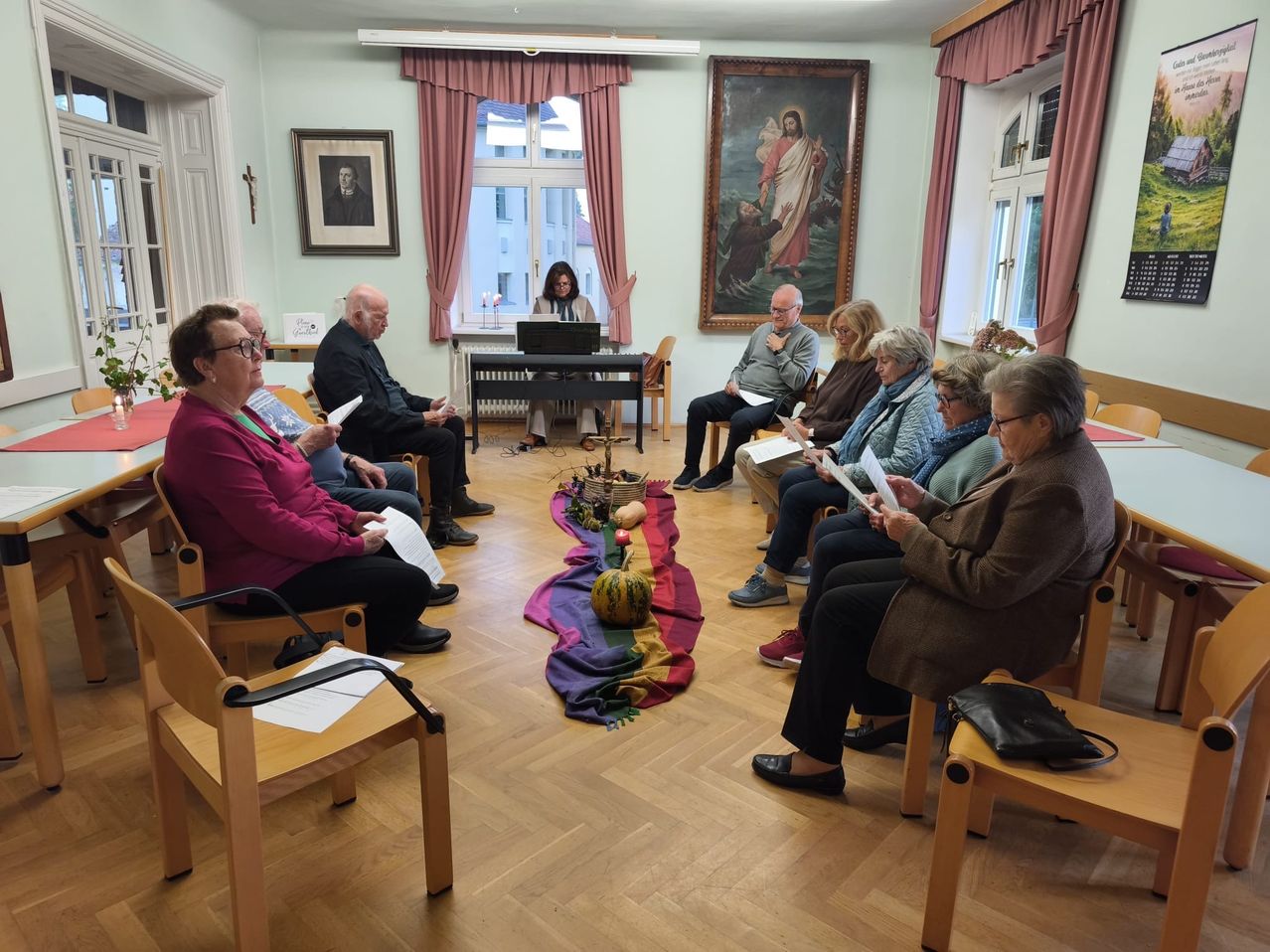 Eine Gruppe von Menschen sitzt im Kreis in einem Raum. Eine Frau steht und liest etwas von einem Laptop. Der Raum hat Holzböden und einige Gemälde an der Wand.
