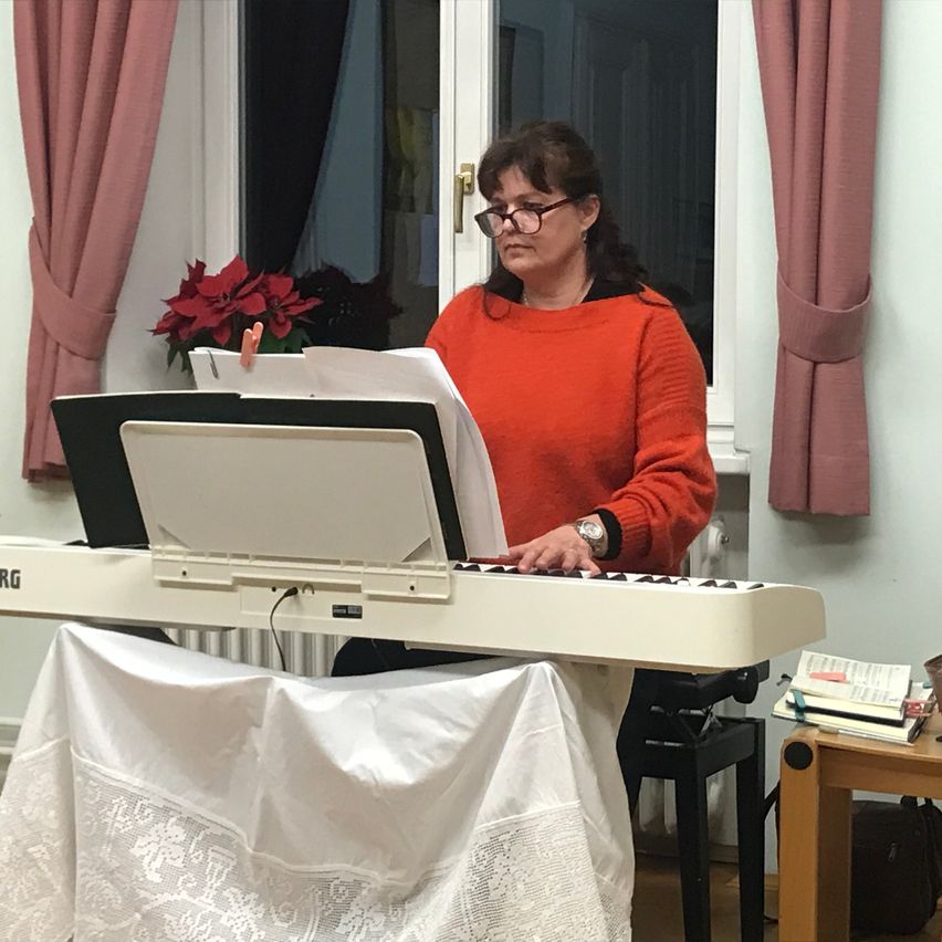 Eine Frau in einem roten Pullover spielt Klavier in einem Raum mit weißer Tischdecke, einem Fenster mit Vorhängen und einer roten Weihnachtsstern.