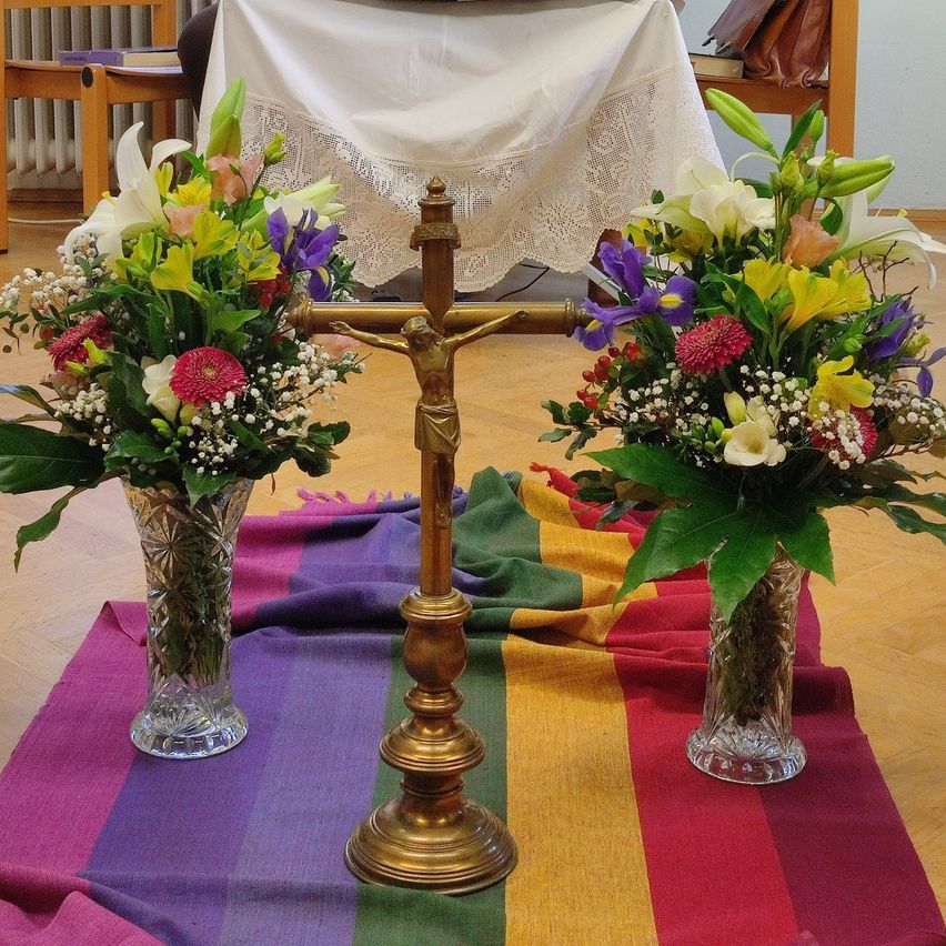 Ein goldenes Kreuz mit einem Kruzifix steht auf einem farbenfrohen Regenbogentuch. Zwei Vasen mit bunten Blumen stehen auf beiden Seiten des Kreuzes. Im Hintergrund bedeckt ein weißes Tuch einen Tisch.