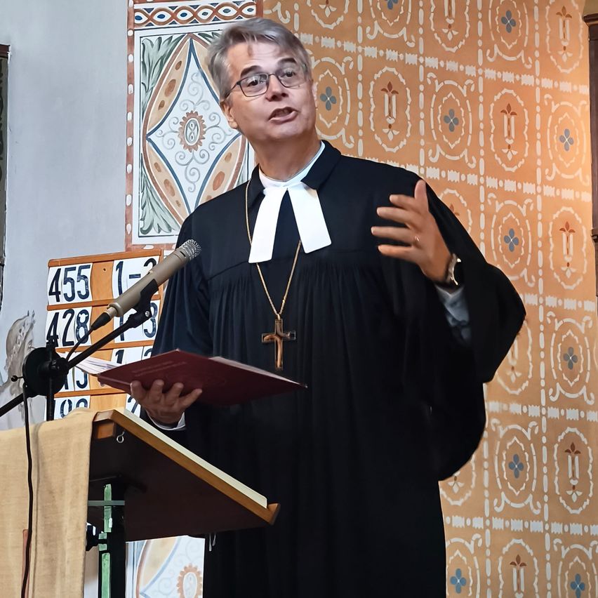 A priest stands at a pulpit, holding a book, and speaking into a microphone. He wears a black robe and a white stole. Behind him is a wall with decorative patterns.