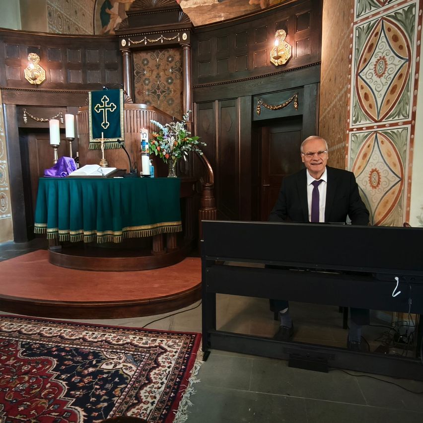 A man in a suit sits at an organ in a church, with an altar and candles behind him.