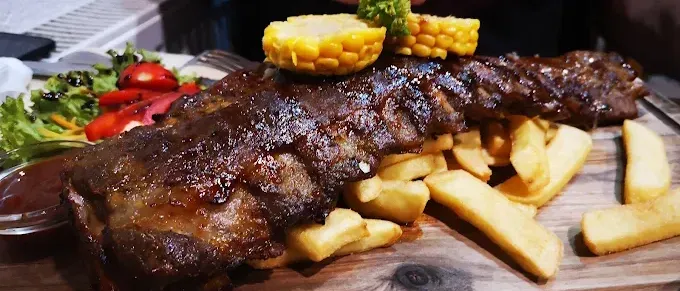 Gegrillte Rippchen mit Maiskolben auf einem Holzschneidebrett, begleitet von Pommes und einem Beilagensalat mit Tomaten und Dressing.