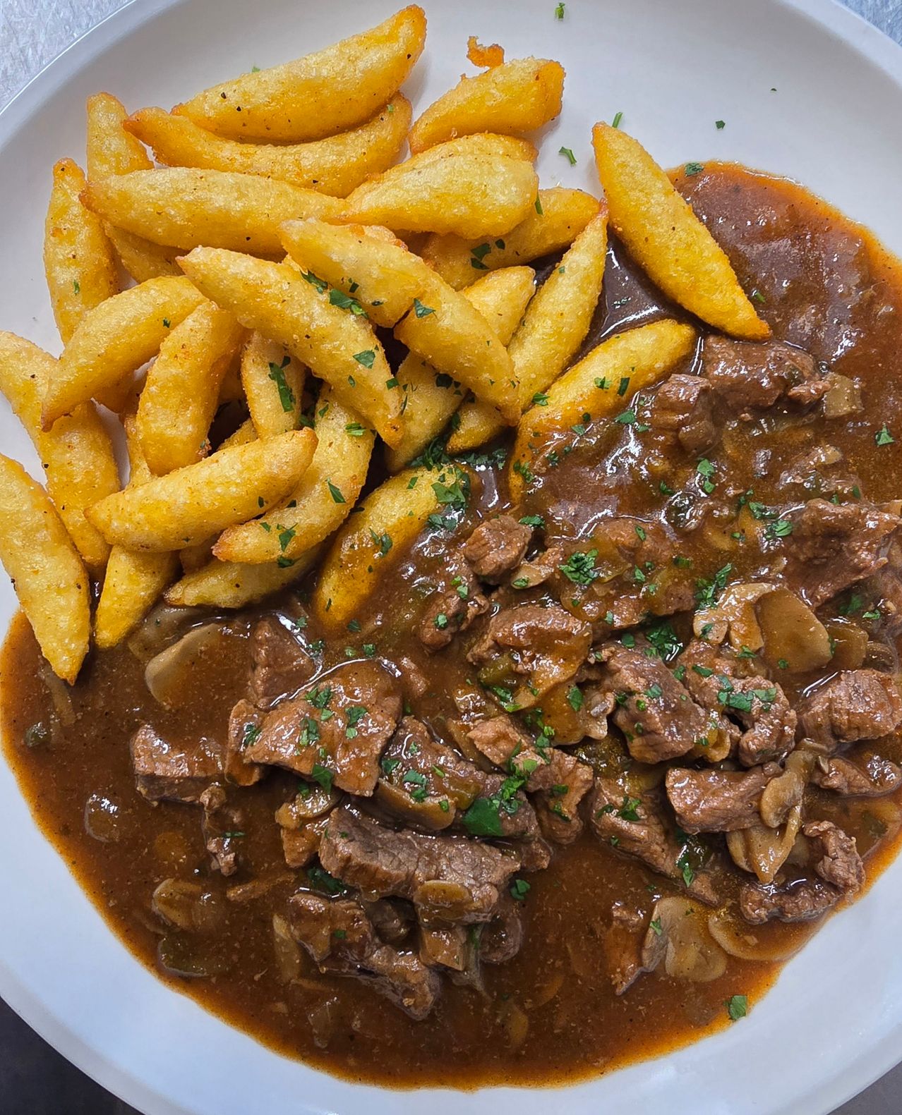 A plate with a serving of beef bourguignon with mushrooms, garnished with parsley, accompanied by a side of golden french fries.