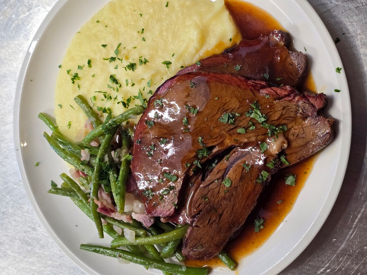 A plate of beef with green beans and mashed potatoes garnished with herbs, topped with a rich gravy.