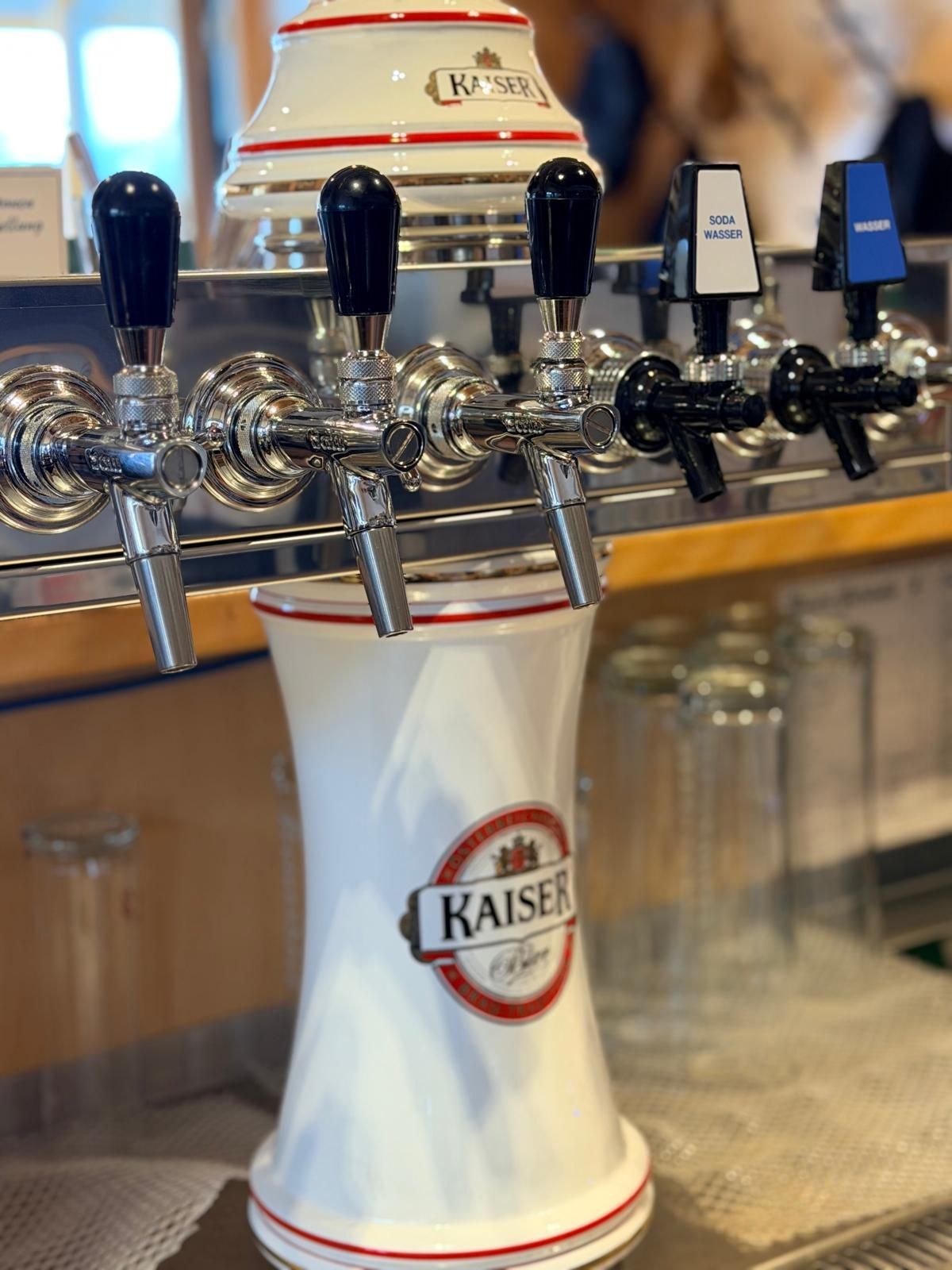 Eine Zapfsäule mit mehreren Bierhähnen und einem Kaiser-Bierkrug in der Mitte. Gläser stehen auf dem Tresen.