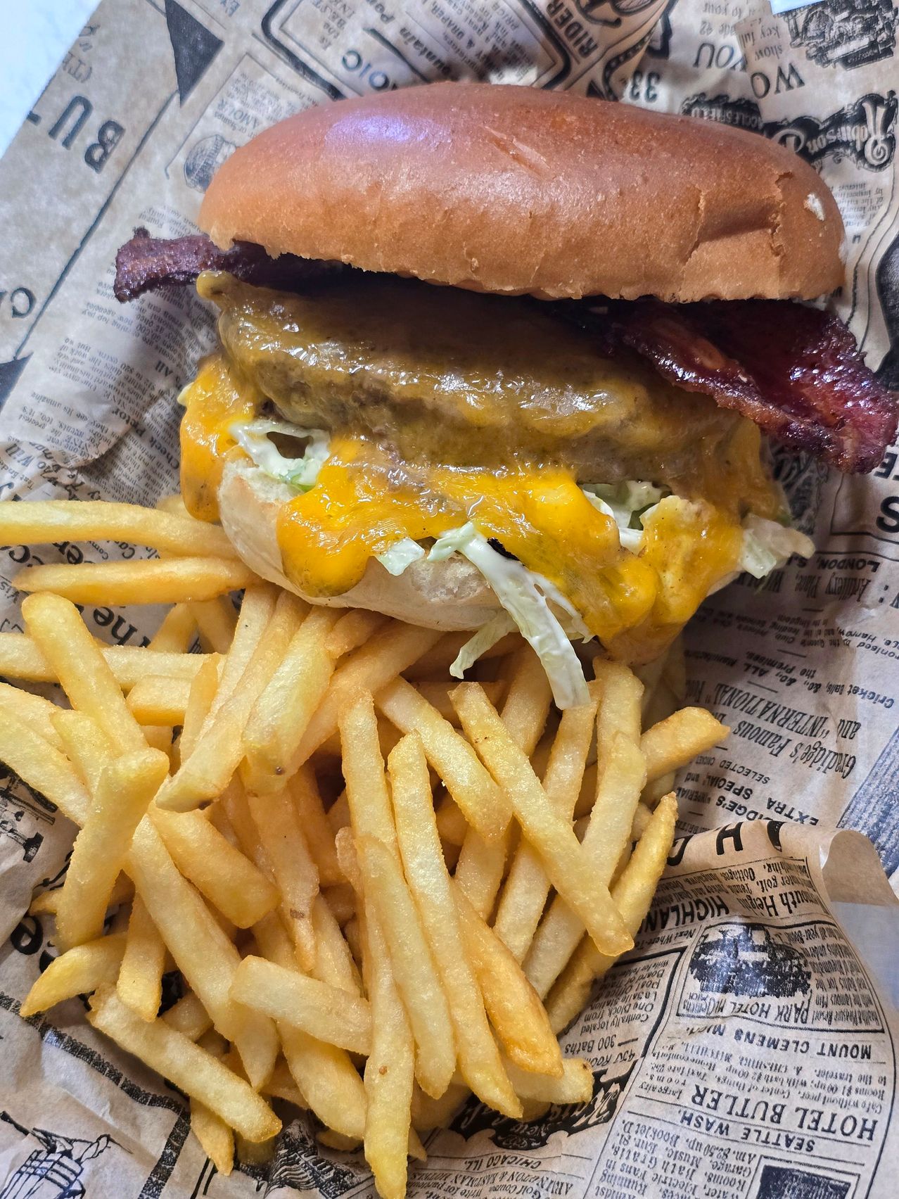 A double cheeseburger with melted cheese, bacon, and lettuce sits atop a bed of golden french fries, all on a newspaper background.