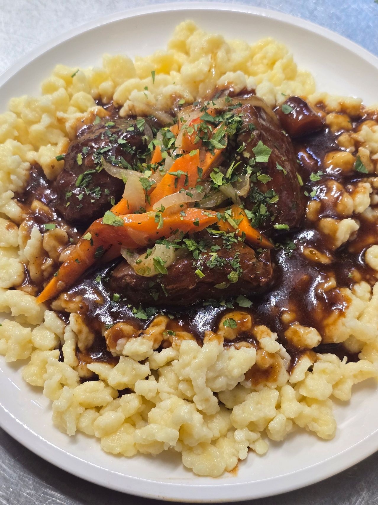 A plate of braised beef with a rich gravy, served over creamy mashed potatoes, and garnished with chopped carrots and parsley.