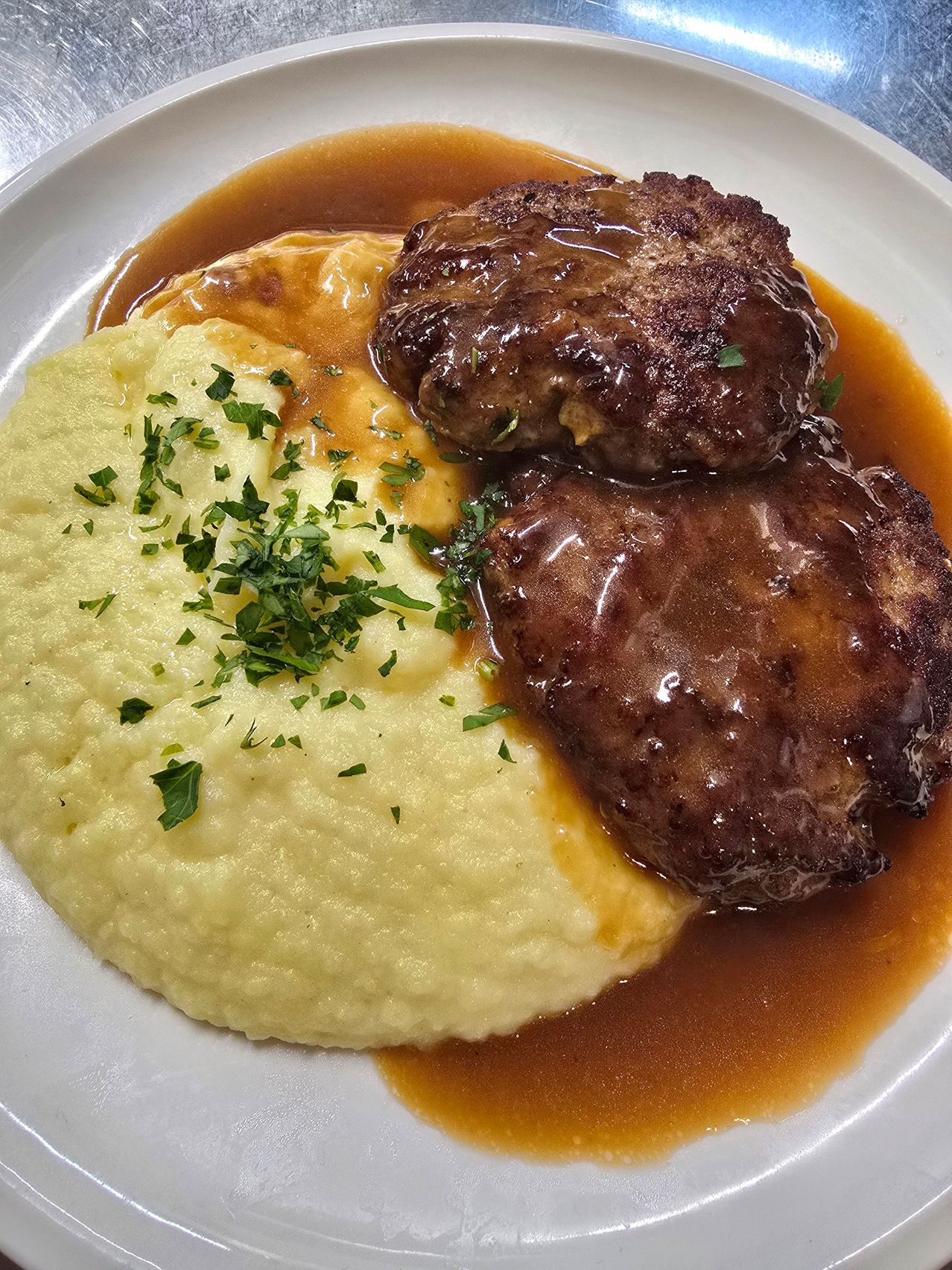 Ein Teller mit zwei in Sauce getauchten Rindfleisch-Patties, verziert mit Kräutern, serviert mit cremigen Kartoffelpüree und brauner Soße.