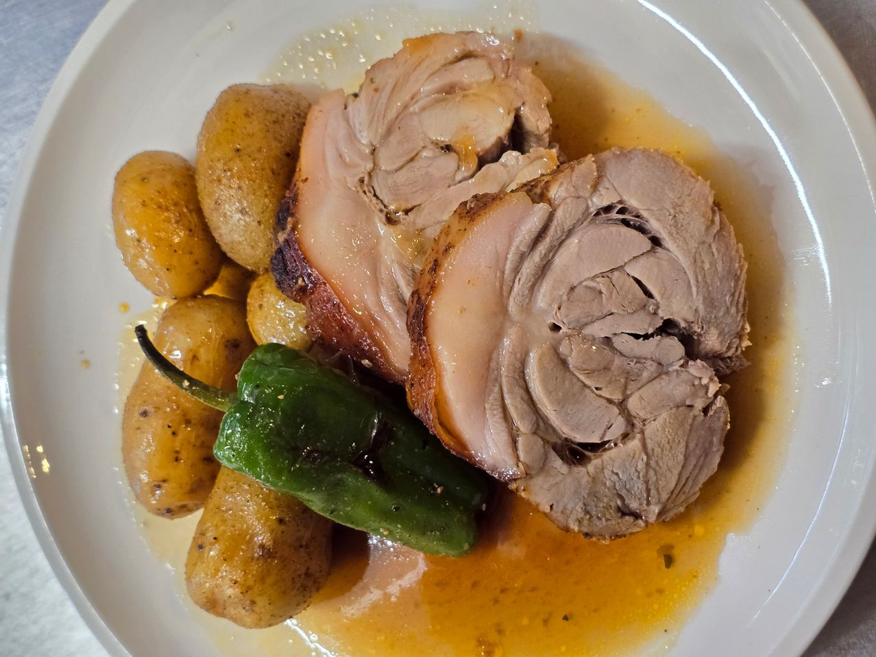 A dish featuring roasted meat, golden potatoes, and a green pepper, all on a white plate. The meat is juicy and slightly crispy.