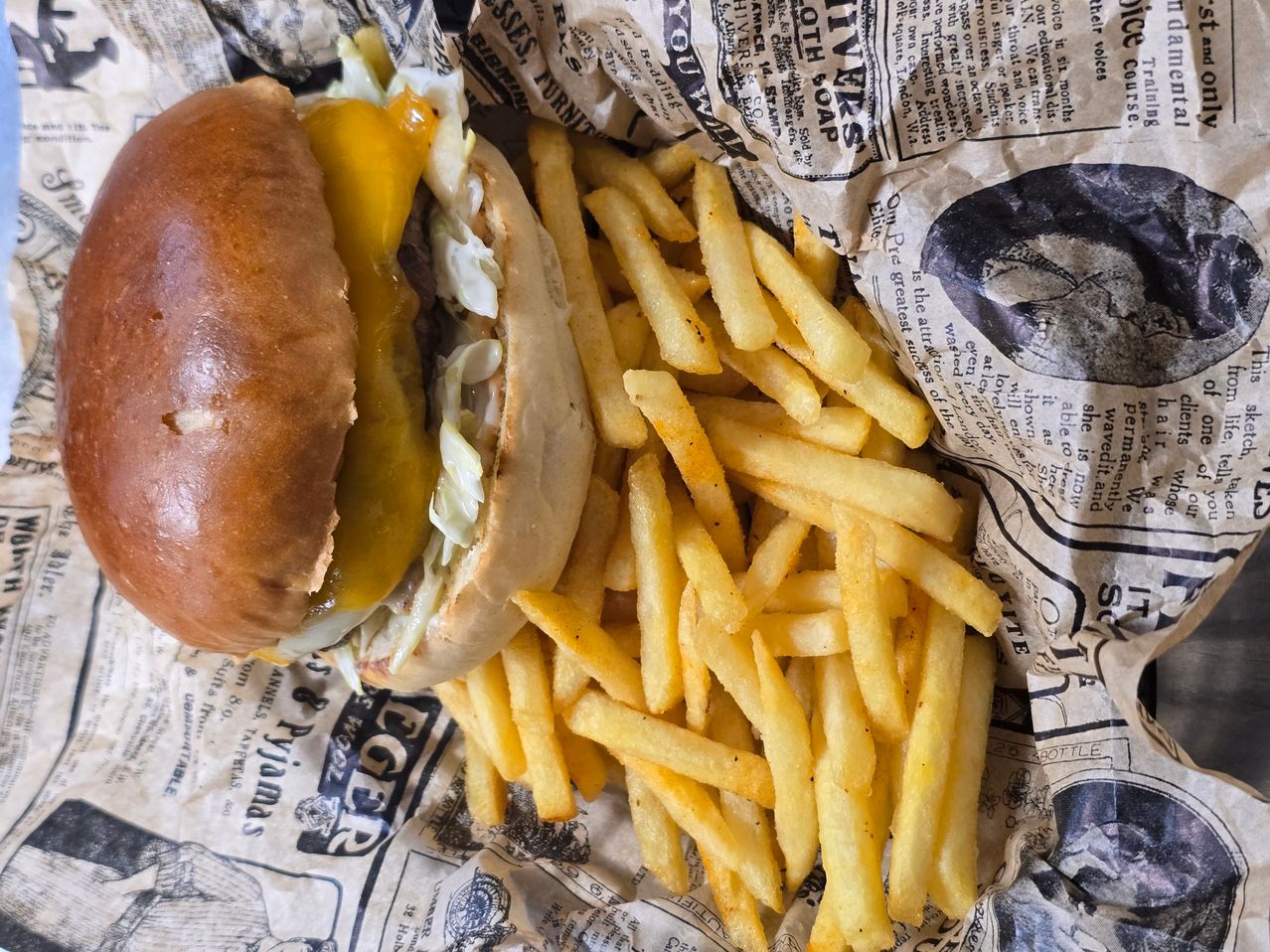 A burger with cheese and fries on a newspaper. The burger is on top of a pile of fries.