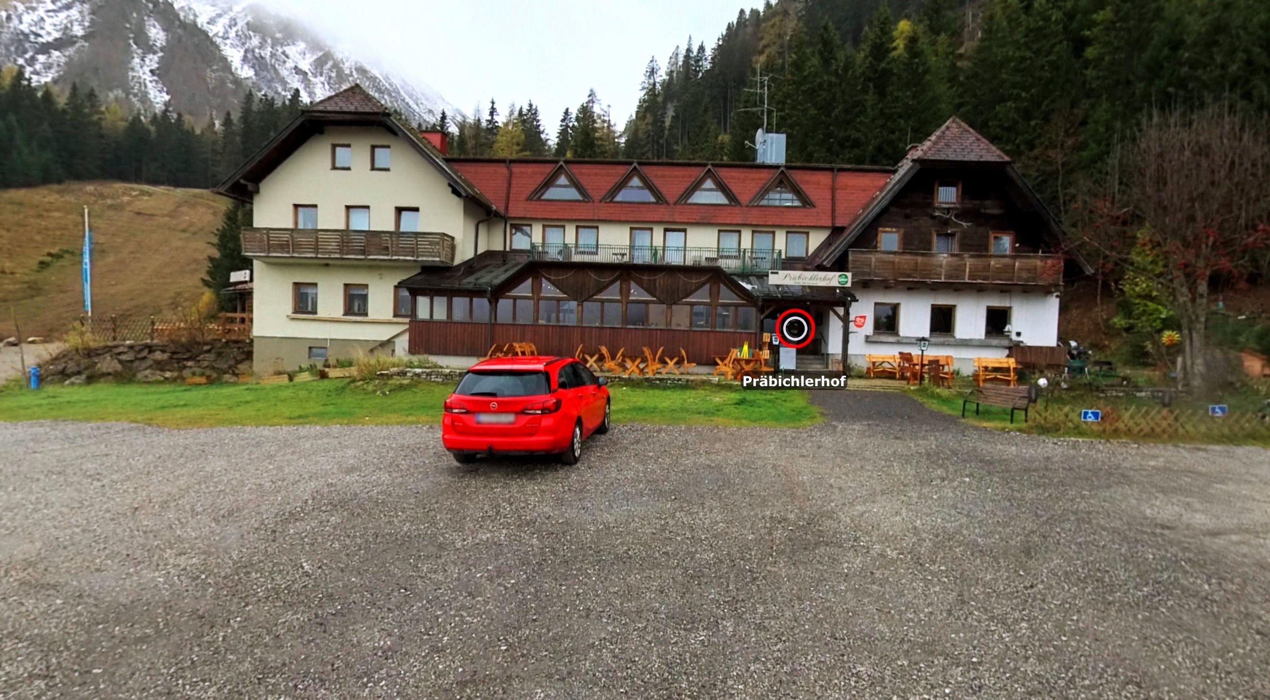 Ein rotes Auto steht vor einem Gebäude namens Prabiclerhof, umgeben von Gartenmöbeln, Rasen und Bergen.