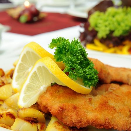 Nahaufnahme eines Tellers mit gebratenem Schnitzel, Zitronenscheiben und Petersiliengarnitur. Im Hintergrund befindet sich eine verschwommene rote Tischdecke und ein weiteres Gericht.