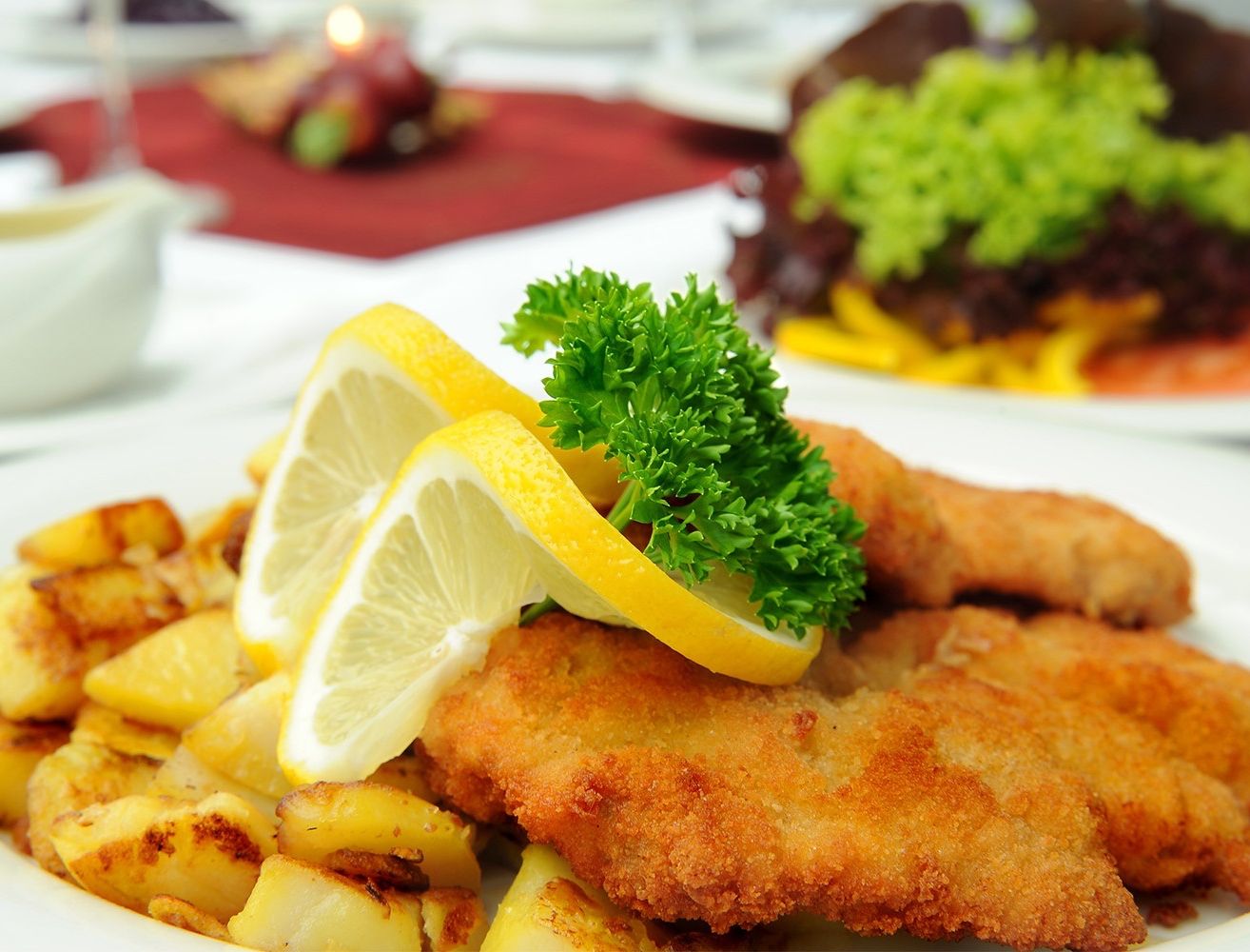Nahaufnahme eines Tellers mit gebratenem Schnitzel, Zitronenscheiben und Petersiliengarnitur. Im Hintergrund befindet sich eine verschwommene rote Tischdecke und ein weiteres Gericht.