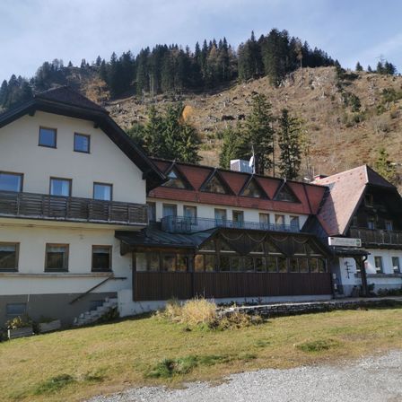 Ein mehrstöckiges Gebäude mit einer braunen Vorderseite steht an einem Hang vor einer Kulisse aus von Kiefern bedeckten Hügeln. Das Gebäude hat einen Balkon mit Geländer, mehrere Fenster und ein Schild, das 'Pension' steht.